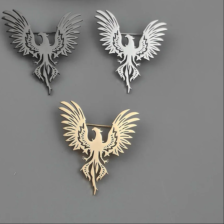 Three phoenix-shaped brooches in gold, silver, and bronze on a gray background.