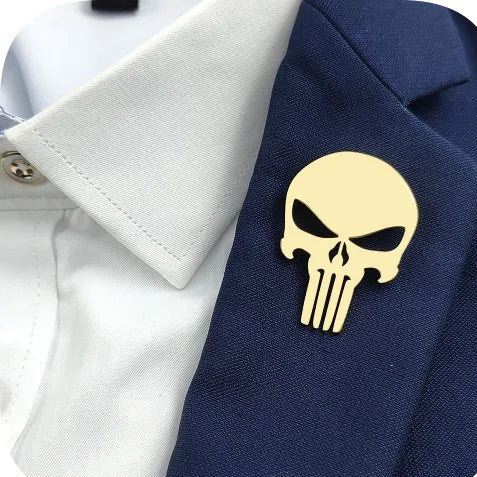 A set of four skull-shaped brooches in gold, white, silver, and black colors, displayed on a grey background.