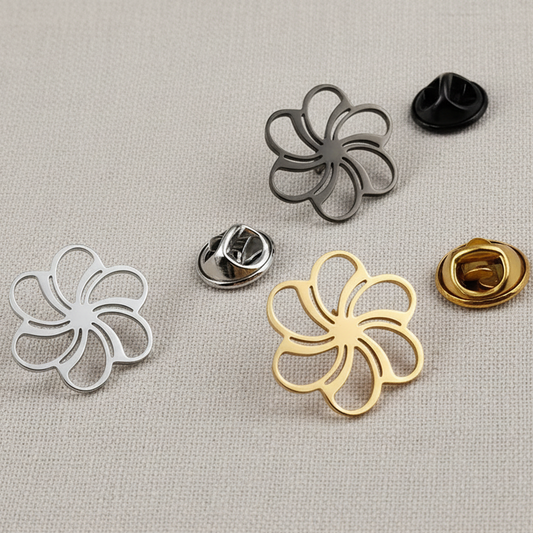 A set of stainless steel brooches in silver, gold, and black colors, with a hollow flower pattern, displayed on a grey surface.