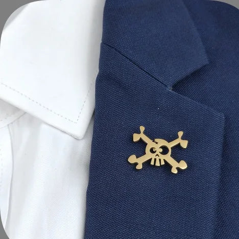 A set of skull-shaped lapel pins in gold and silver colors displayed on a grey background, with one pin attached to a blue fabric possibly representing a suit.