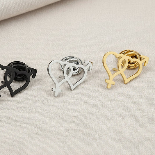 A set of metal brooches in the shape of a heart, displayed in gold, silver, and rose gold colors, placed on a wooden surface.