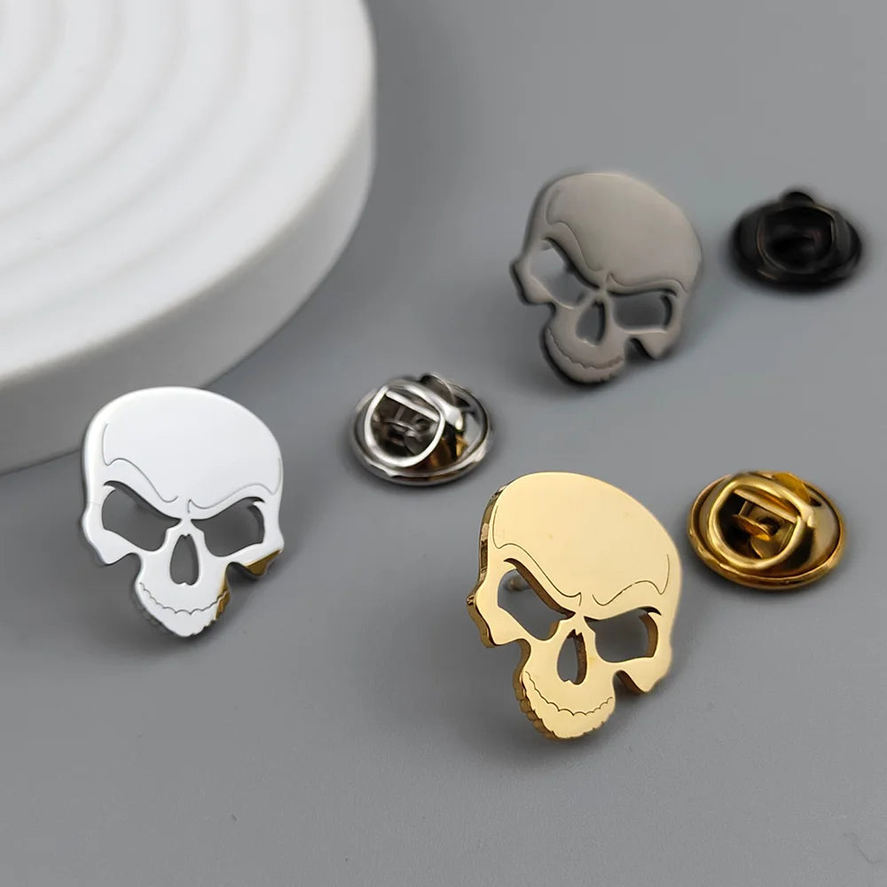 A set of stainless steel skull brooches in black, white, and gold colors, displayed on a grey surface.
